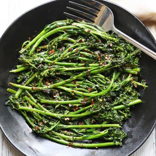 Grilled broccolini served on a dark plate with a fork.
