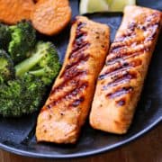 Grilled salmon served on a dark plate with broccoli and sweet potatoes.