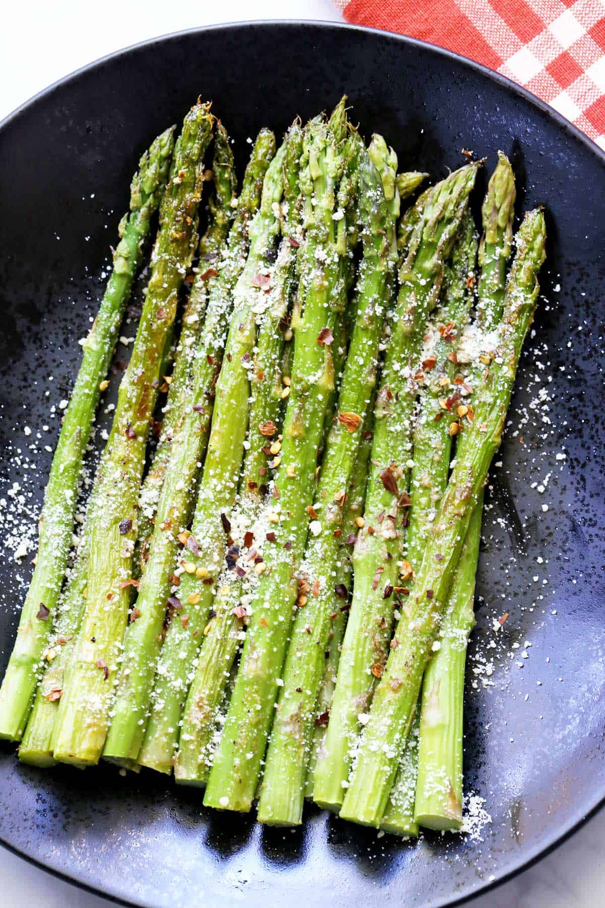 Roasted asparagus is served on a black plate, sprinkled with parmesan and red pepper flakes. 