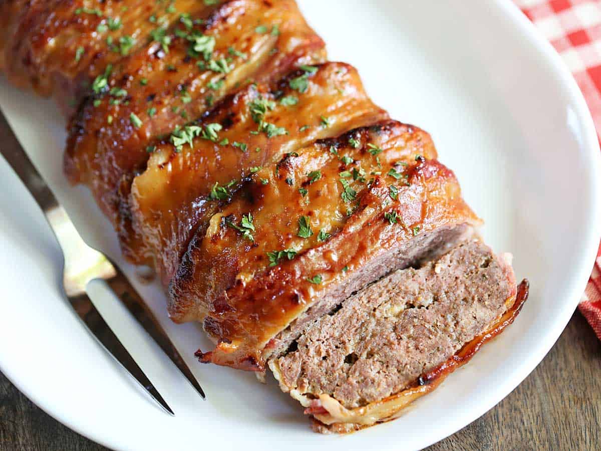 A sliced bacon-wrapped meatloaf is served on a white platter with a serving fork. 