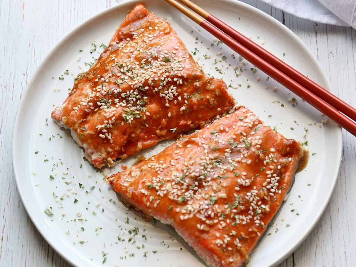 Two pieces of teriyaki salmon served on a white plate with chopsticks. 