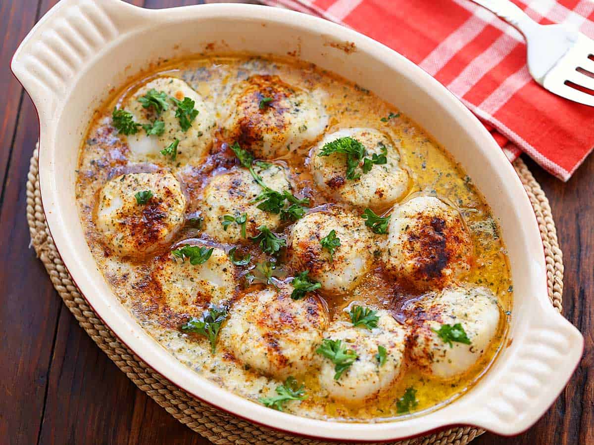 Baked scallops topped with chopped parsley, served in an oval baking dish. 