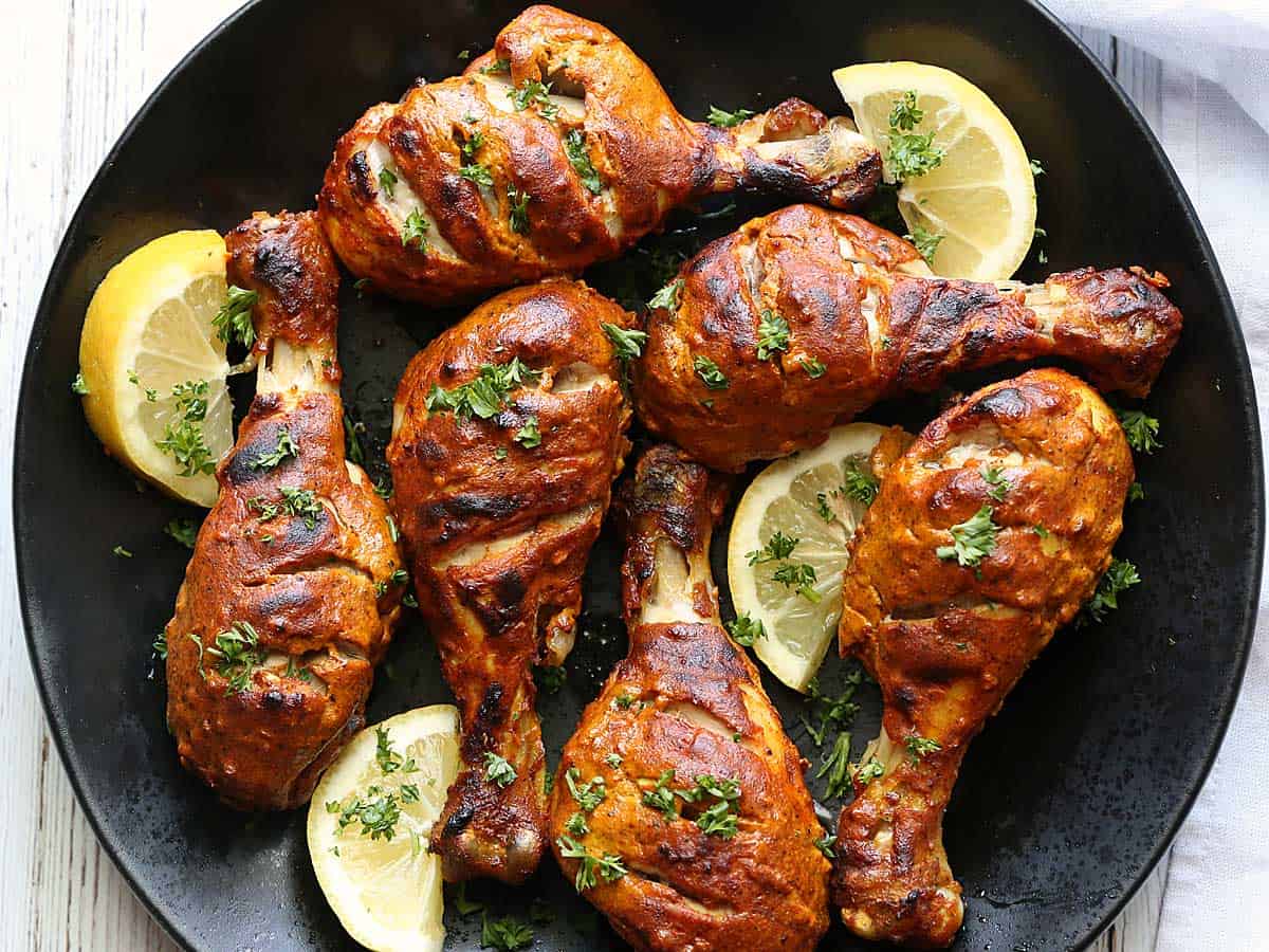 Oven-baked tandoori chicken garnished with parsley, served on a black plate with lemon slices. 