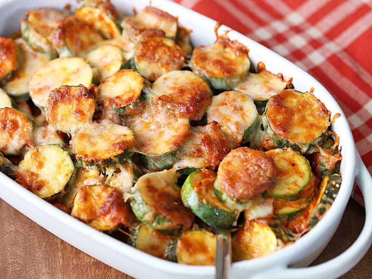 A zucchini casserole is served in a white baking dish with a red napkin.