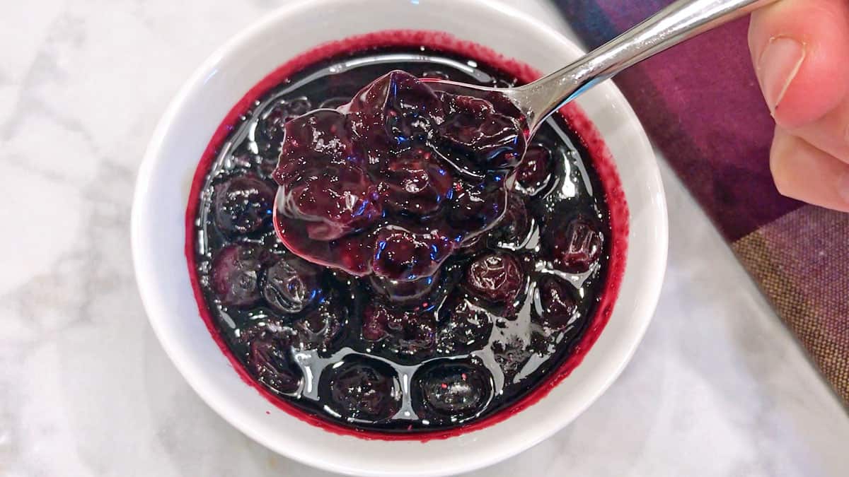 Blueberry compote is ready and served in a white bowl with a spoon. 