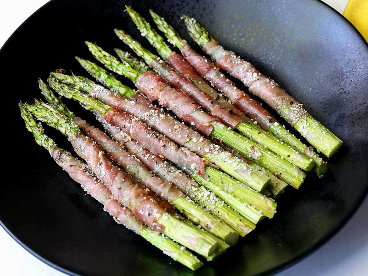 Prosciutto-wrapped asparagus is served on a black plate. 