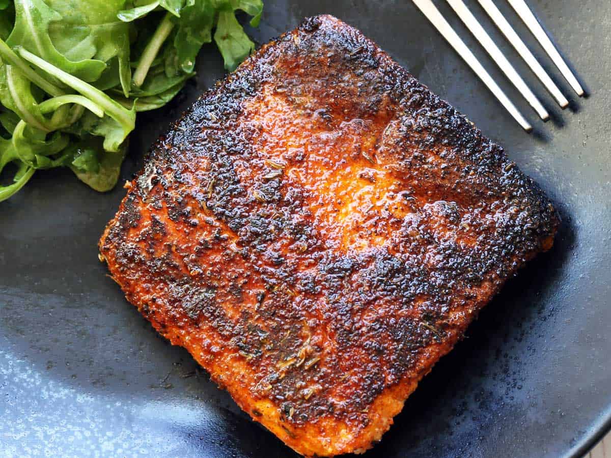 Blackened halibut served on a dark dinner plate with a fork and a side of arugula. 