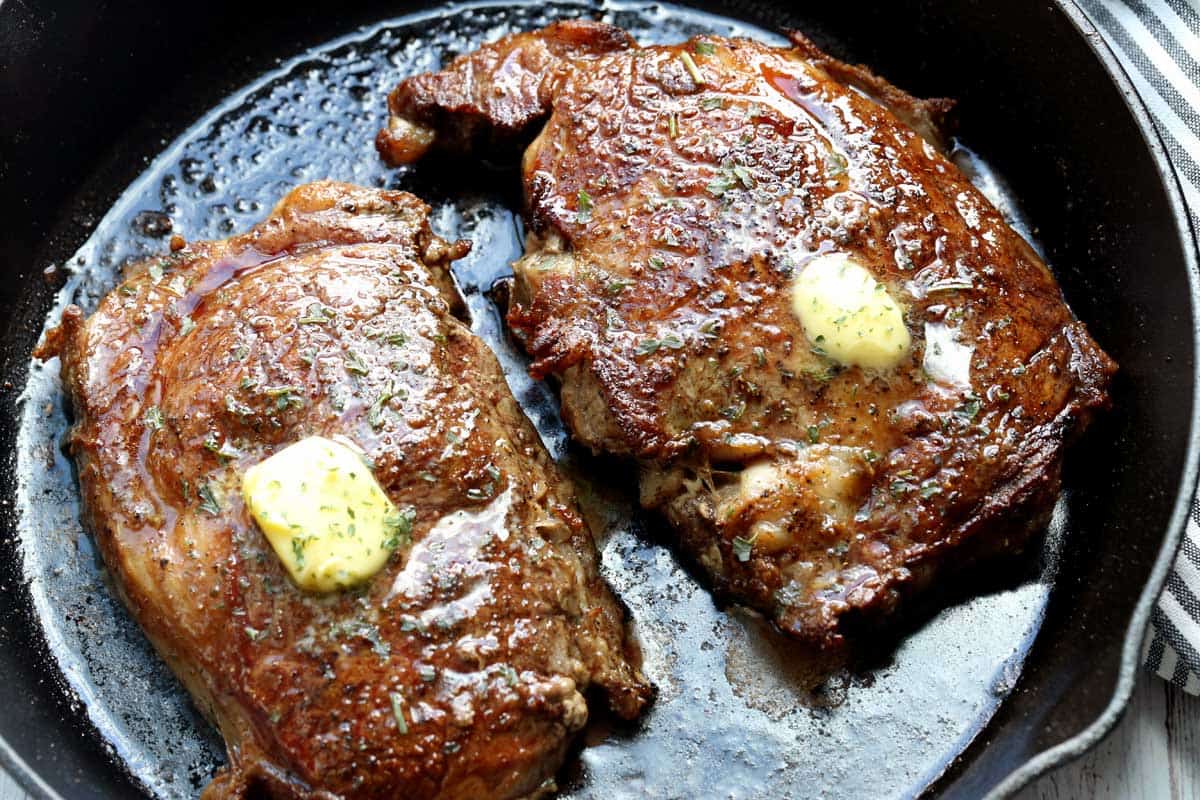 Two boneless ribeye steaks in a cast-iron skillet, topped with butter.