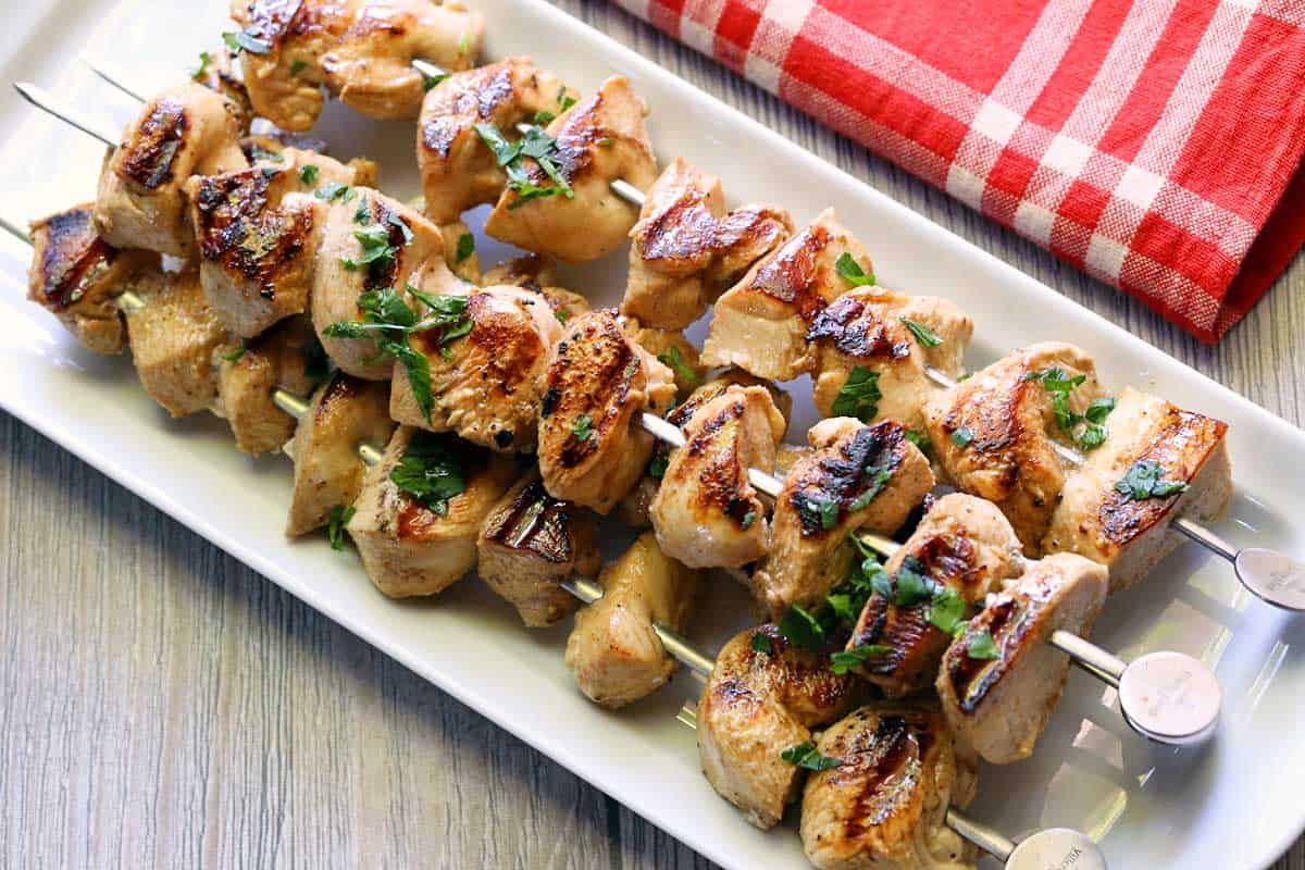 Grilled chicken skewers are served on a white platter with a red napkin. 