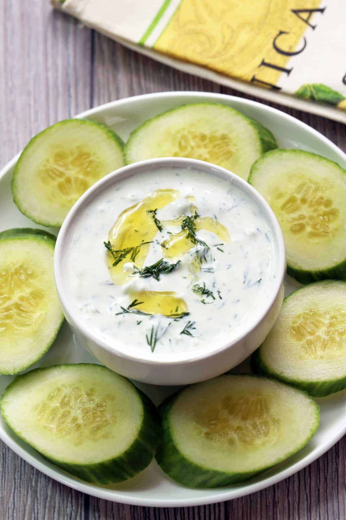 Tzatziki sauce is served in a white bowl with cucumber slices. 