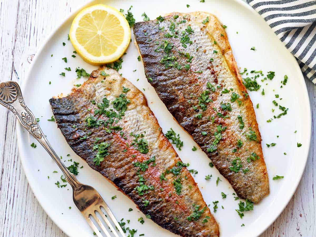 Two fillets of rainbow trout served on a white plate with a slice of lemon. 