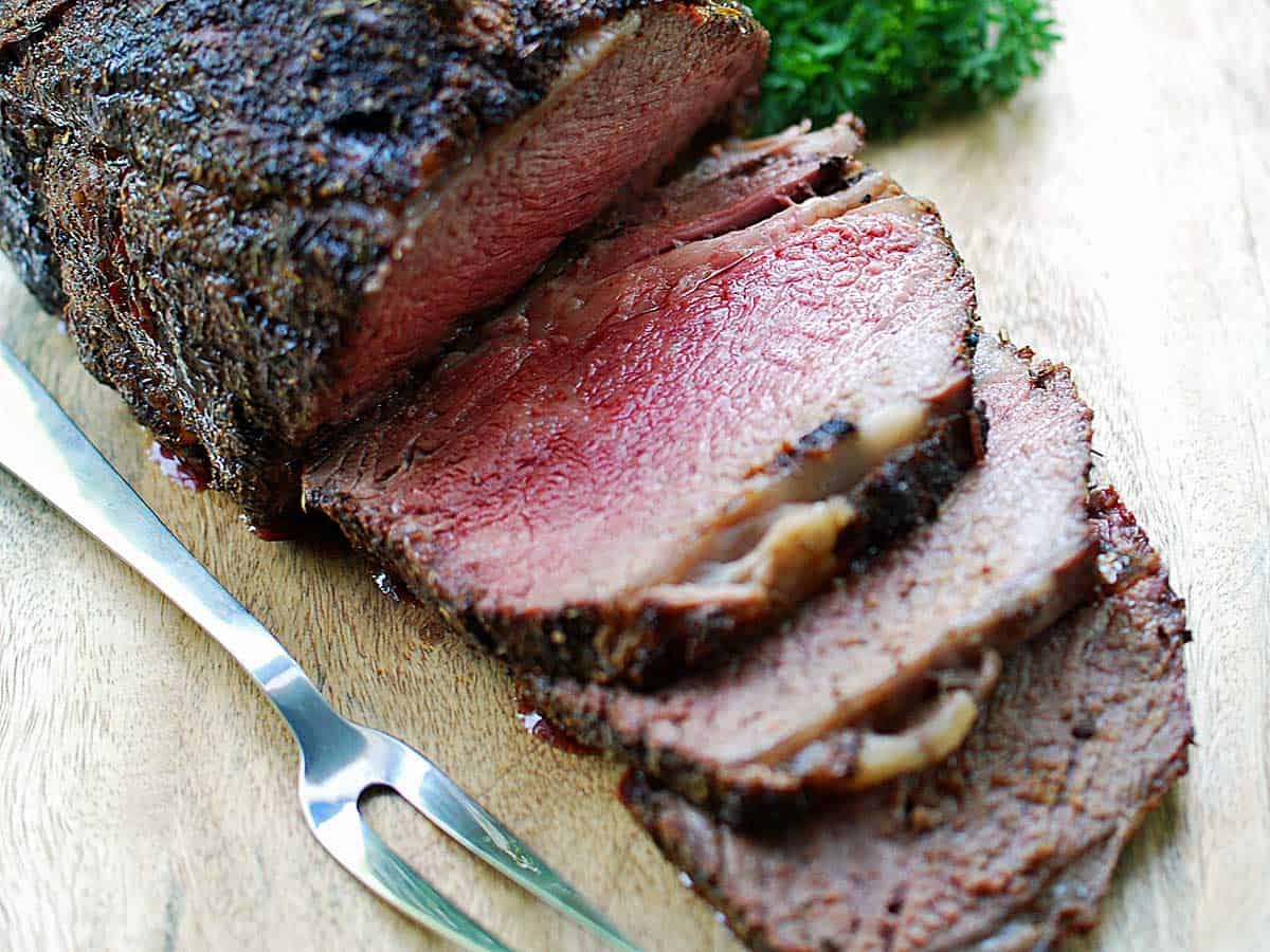 A ribeye roast is served on a wooden cutting board with a serving fork. 