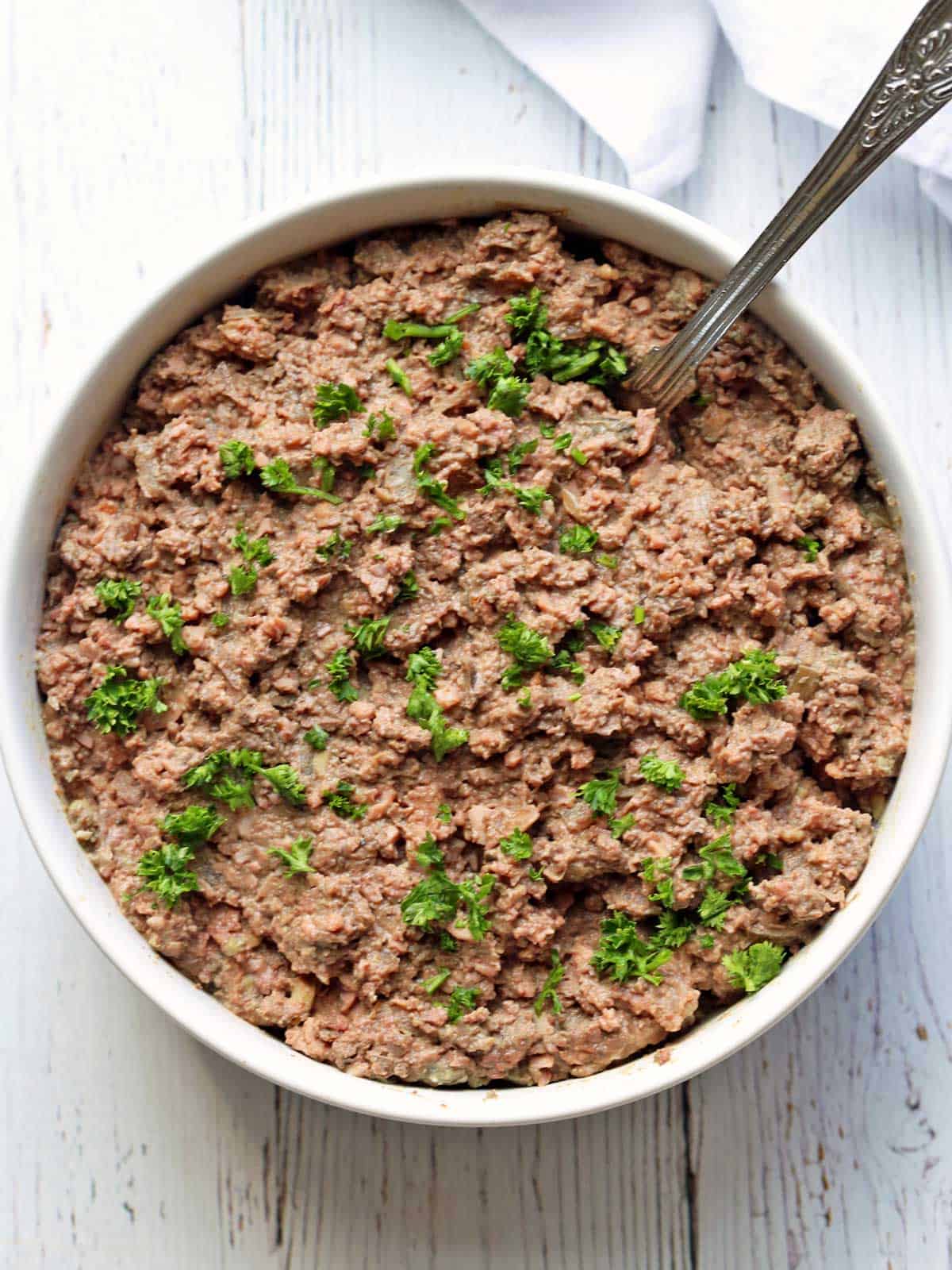 Chopped liver is served in a white bowl with a spoon, topped with parsley.