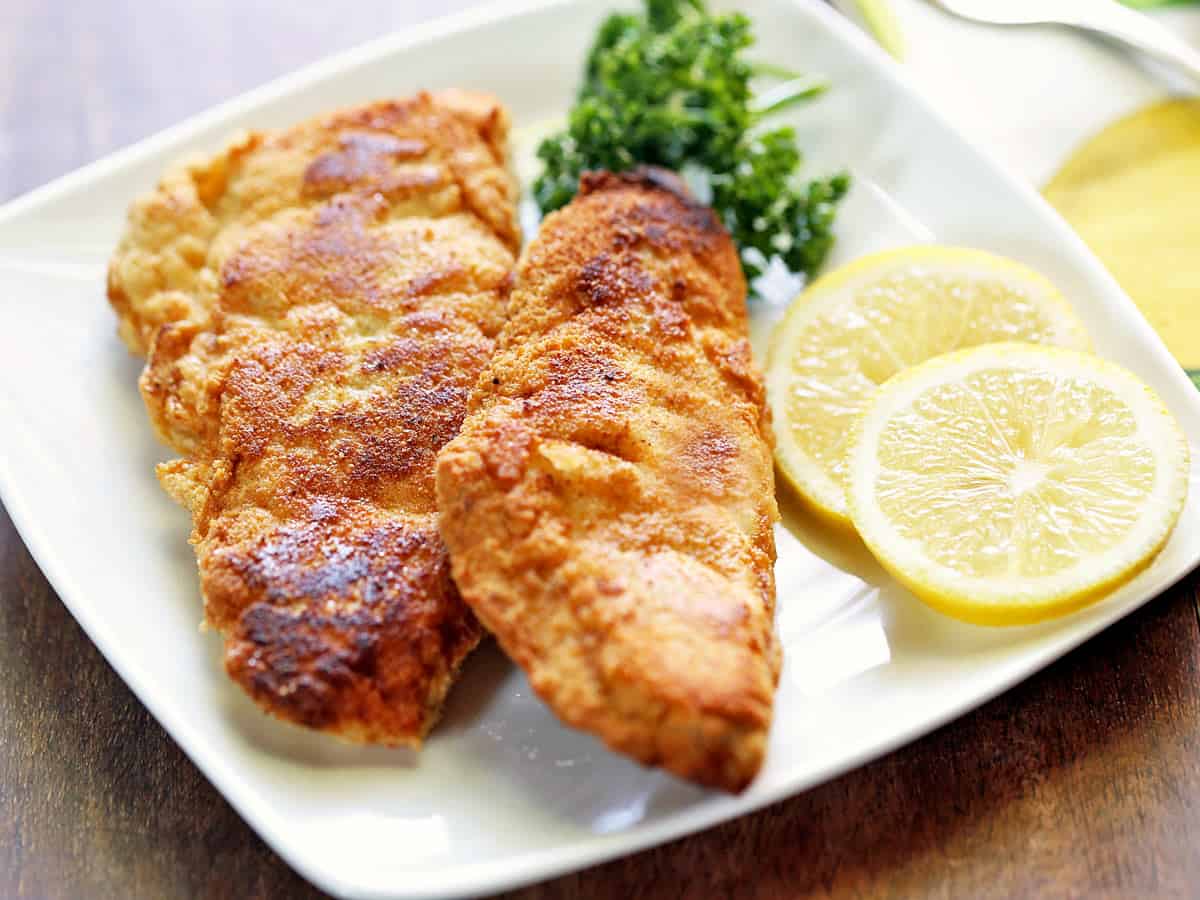 Two pieces of keto fried fish served on a white plate with lemon and parsley. 