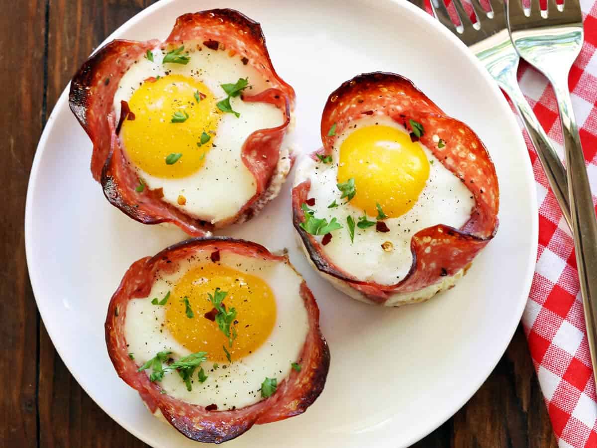 Three salami and eggs cups served on a white plate with a napkin. 