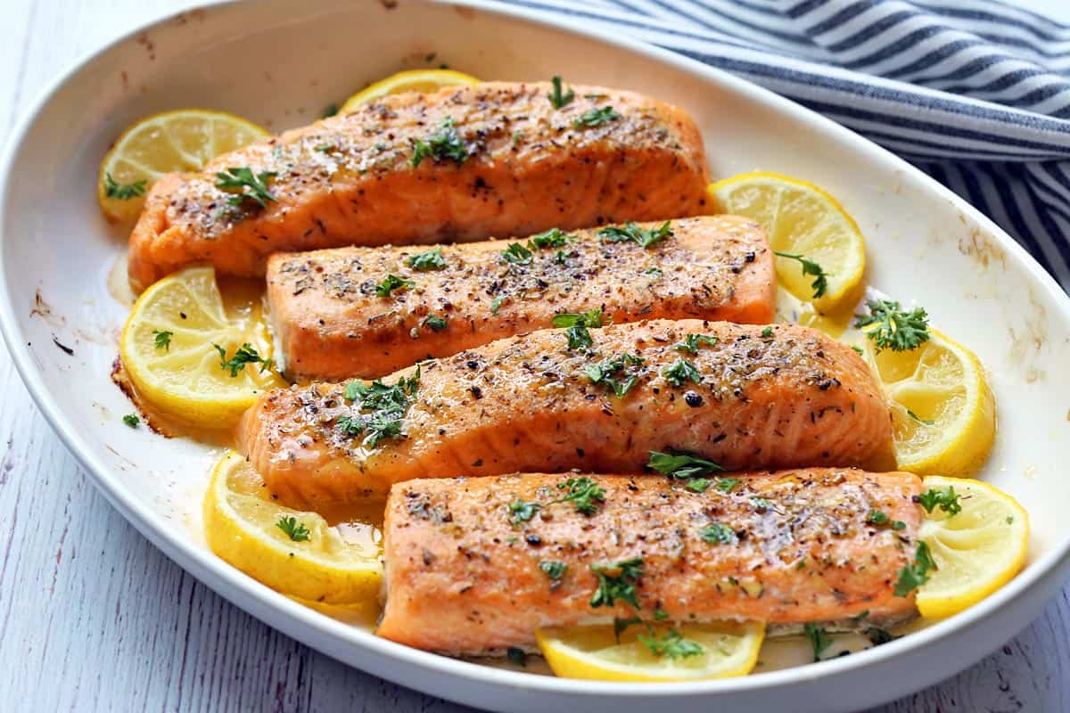 Four salmon fillets served in a white baking dish with lemon slices and chopped parsley. 