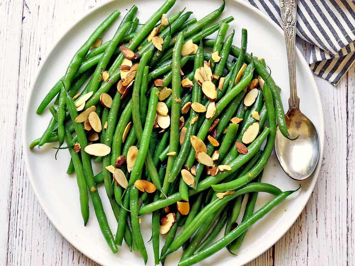 Green beans almondine served on a white plate with a silver spoon and a striped napkin. 