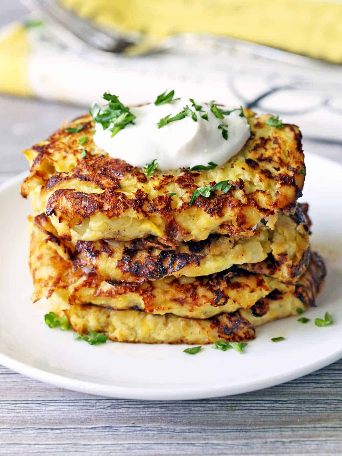 Yellow squash fritters stacked on a white plate and topped with sour cream. 