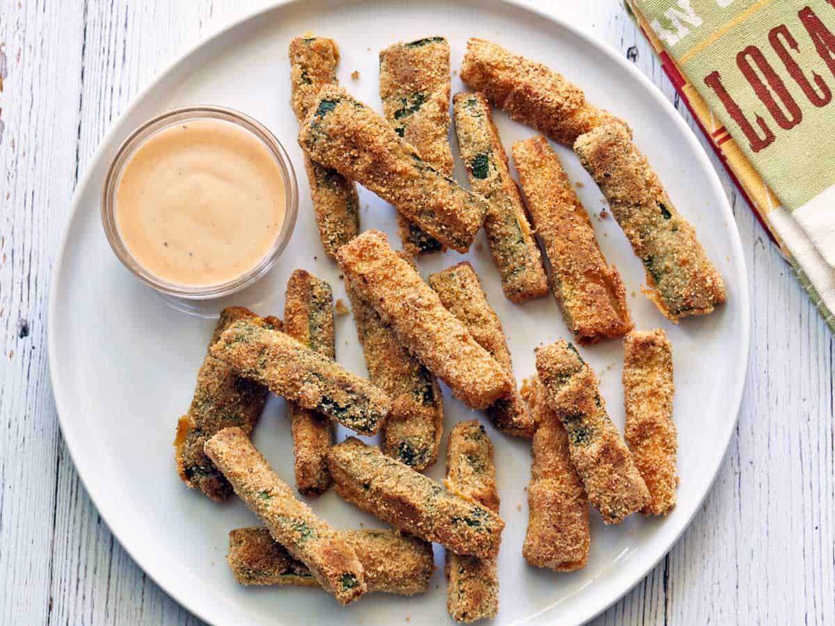 Zucchini fries are served on a white plate with a dipping sauce. 