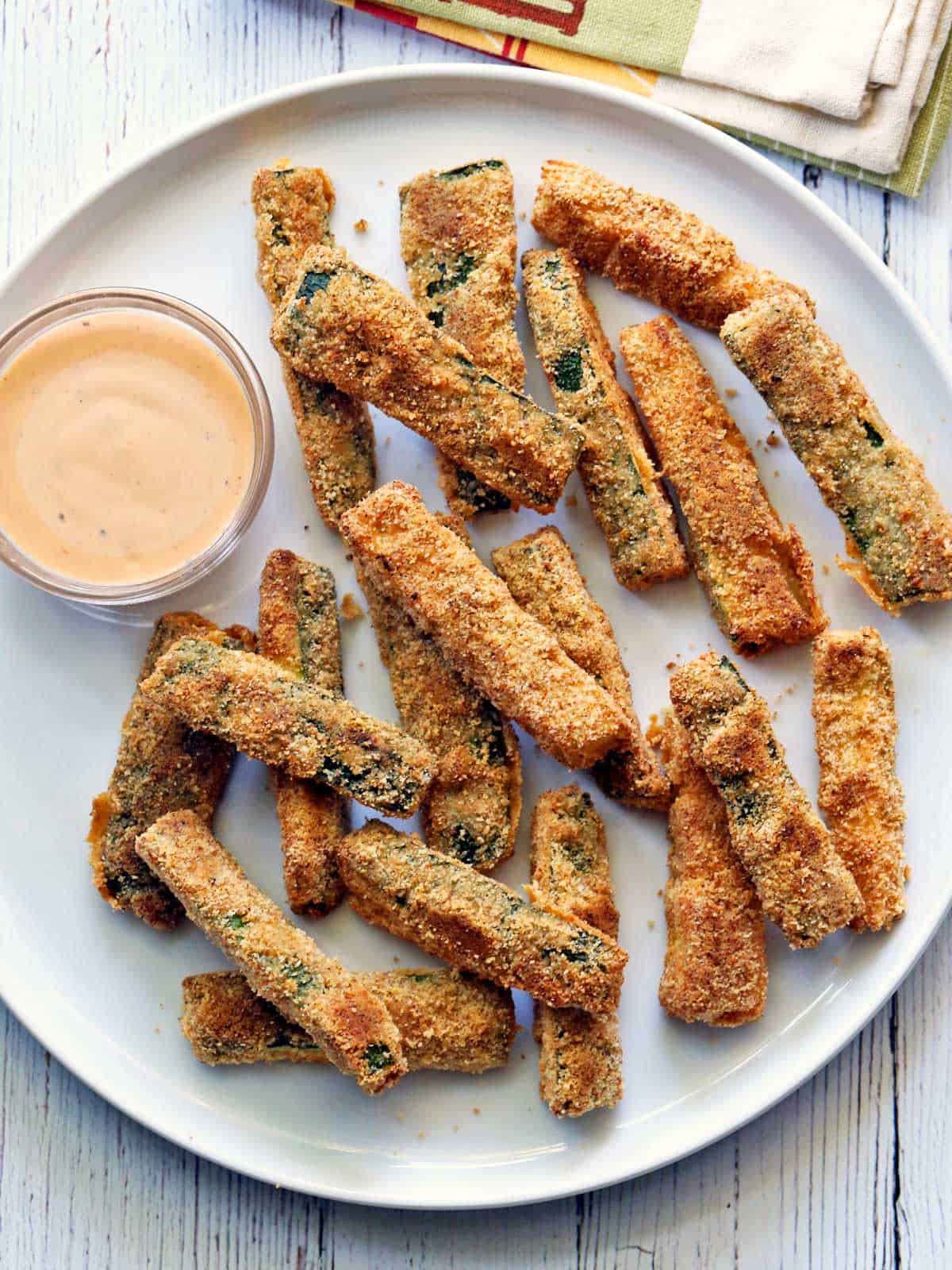 Zucchini fries are served on a white plate with a dipping sauce. 