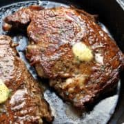 Two ribeye steaks are topped with butter in a cast-iron skillet.