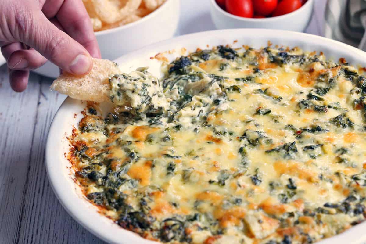 A closeup of a spinach artichoke dip being scooped out with pork rinds. 
