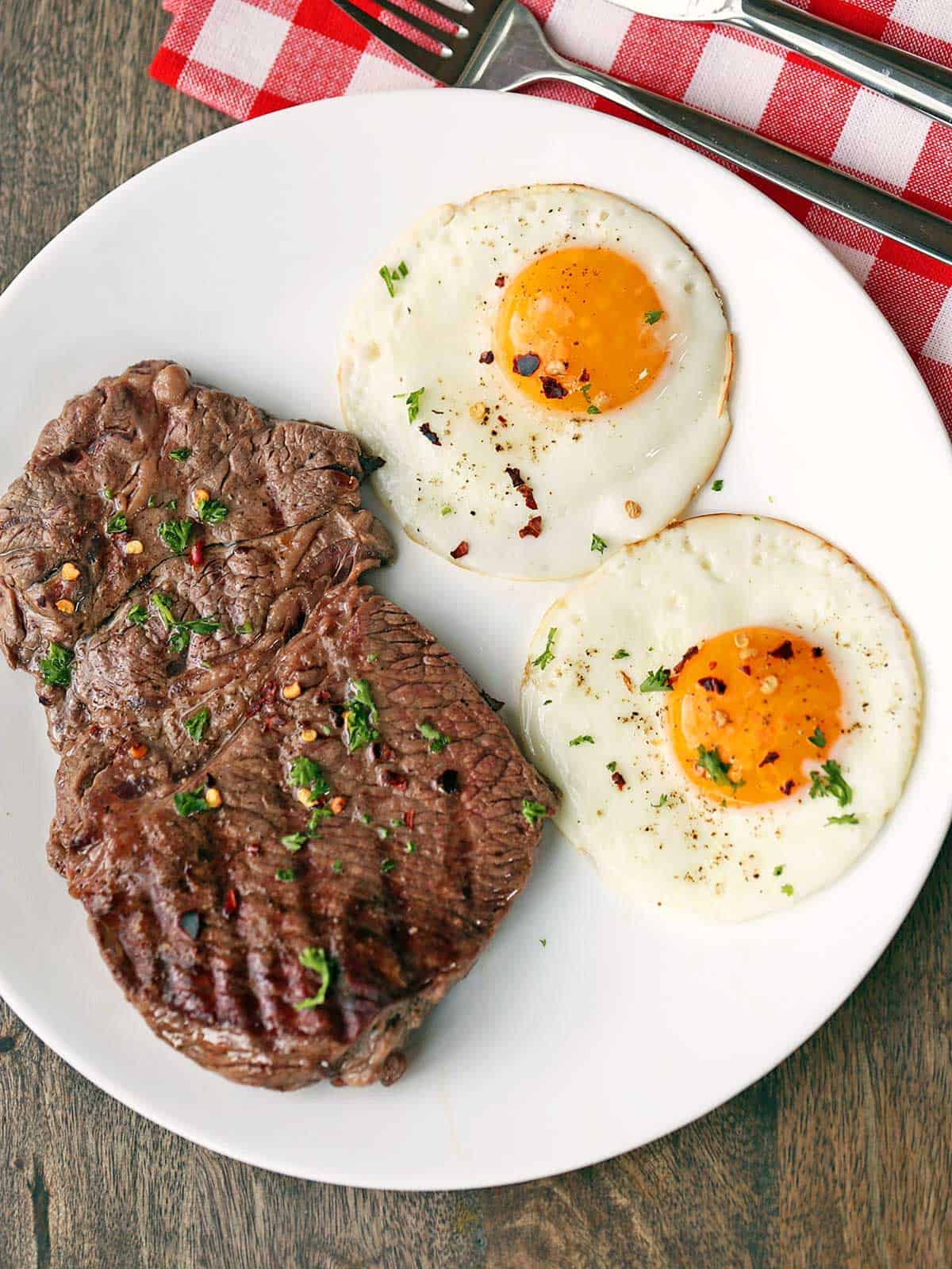 Steak and sunny side up eggs served on a plate with a knife and a fork. 