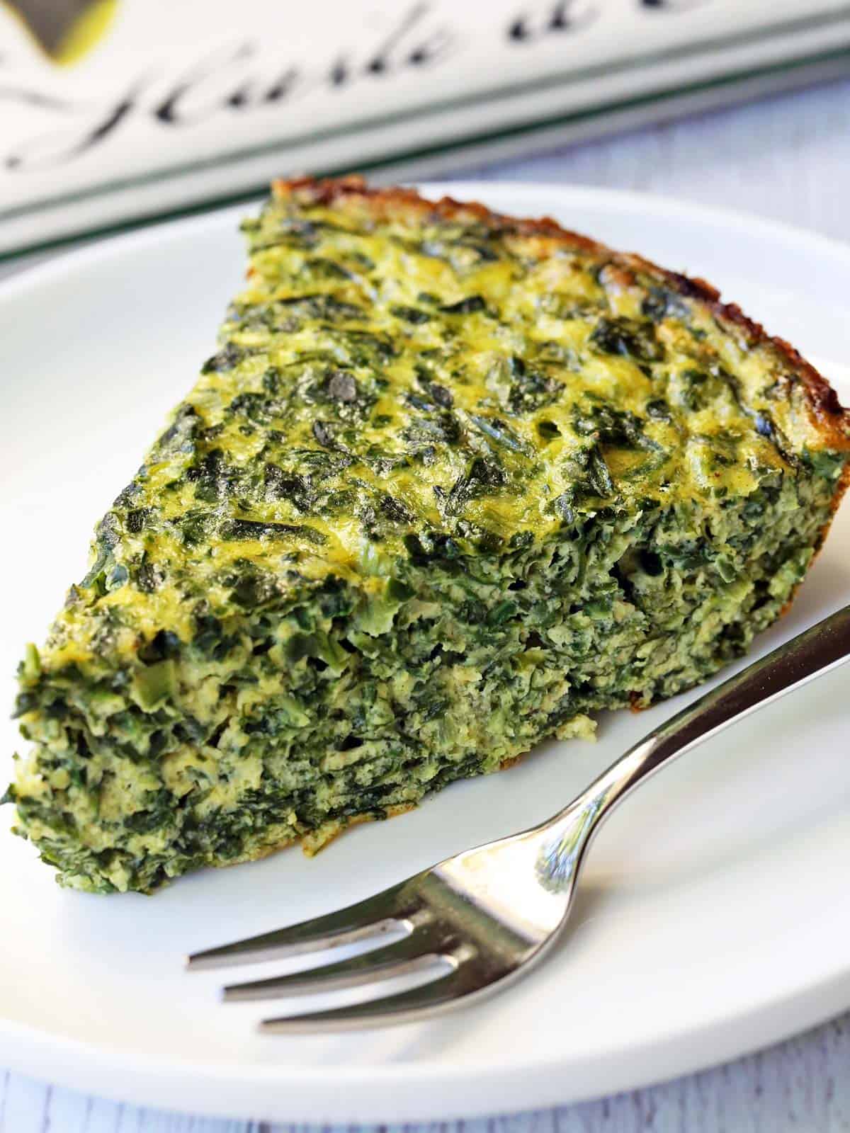 A slice of a crustless spinach quiche served on a white plate with a fork. 