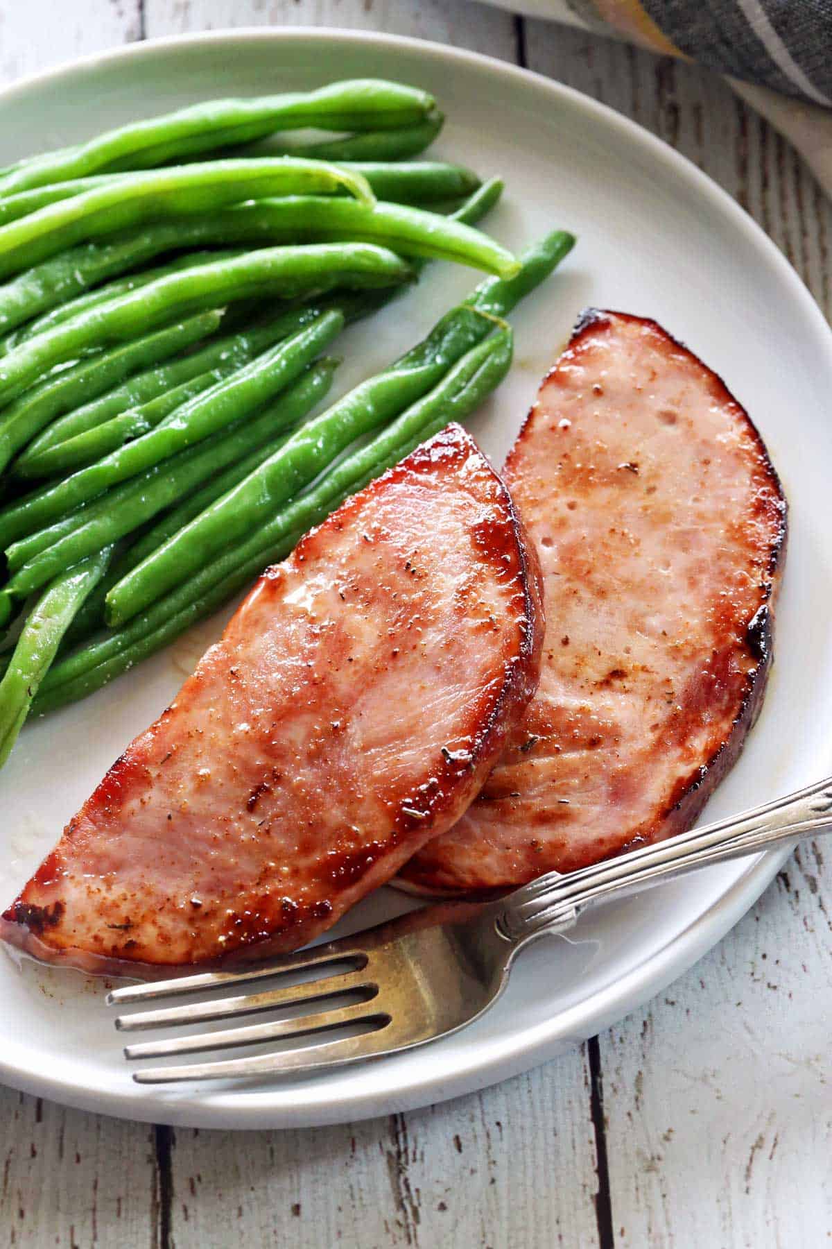 Ham steaks served on a white plate with a fork and a side of green beans.  