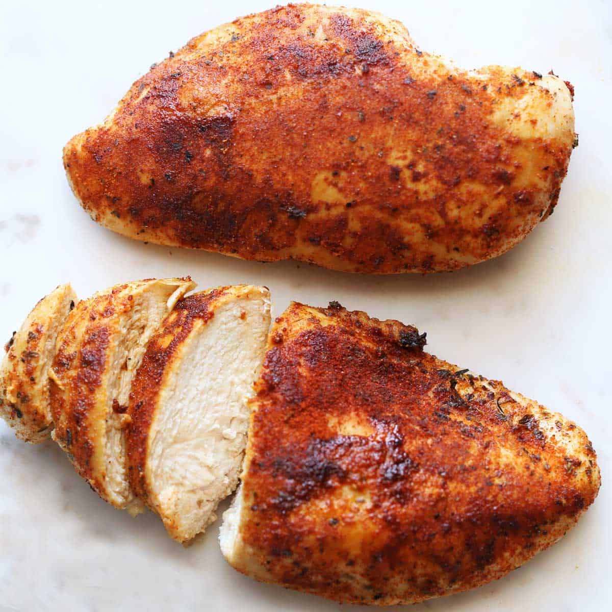 Two baked chicken breasts, well-seasoned, on a white cutting board. 
