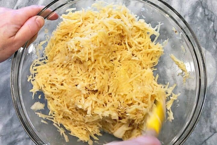 Mixing shredded potatoes, chopped onions, eggs, and spices in a bowl.