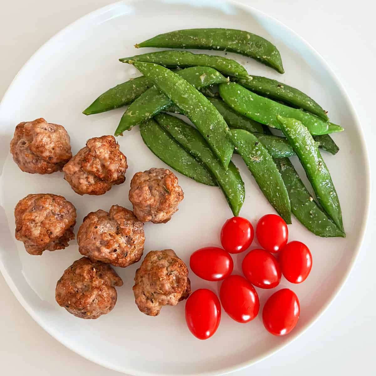 sugar snap peas are served on a white plate with pork meatballs and cherry tomatoes.
