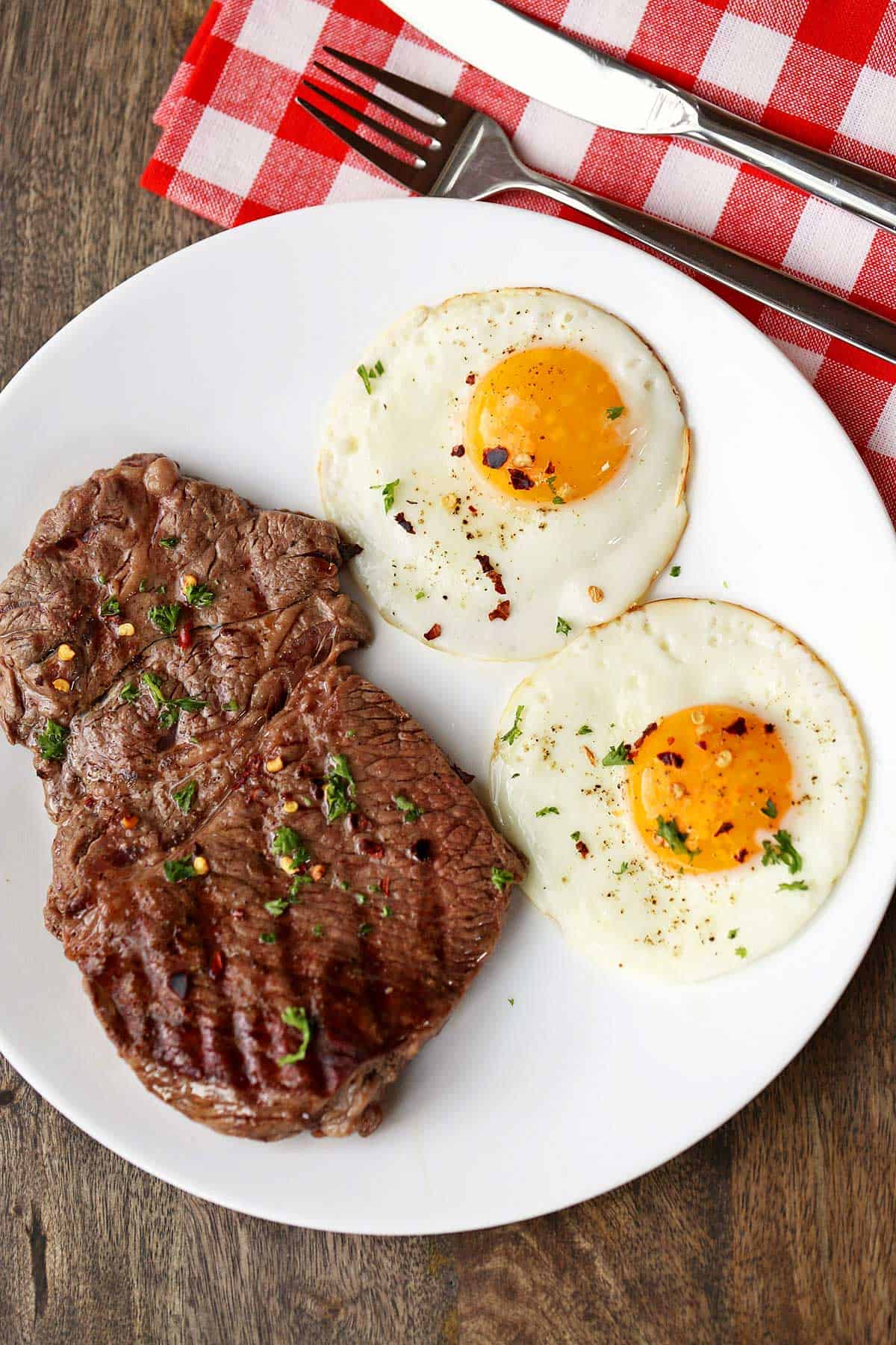 Steak and eggs are served on a white plate with a knife and fork.