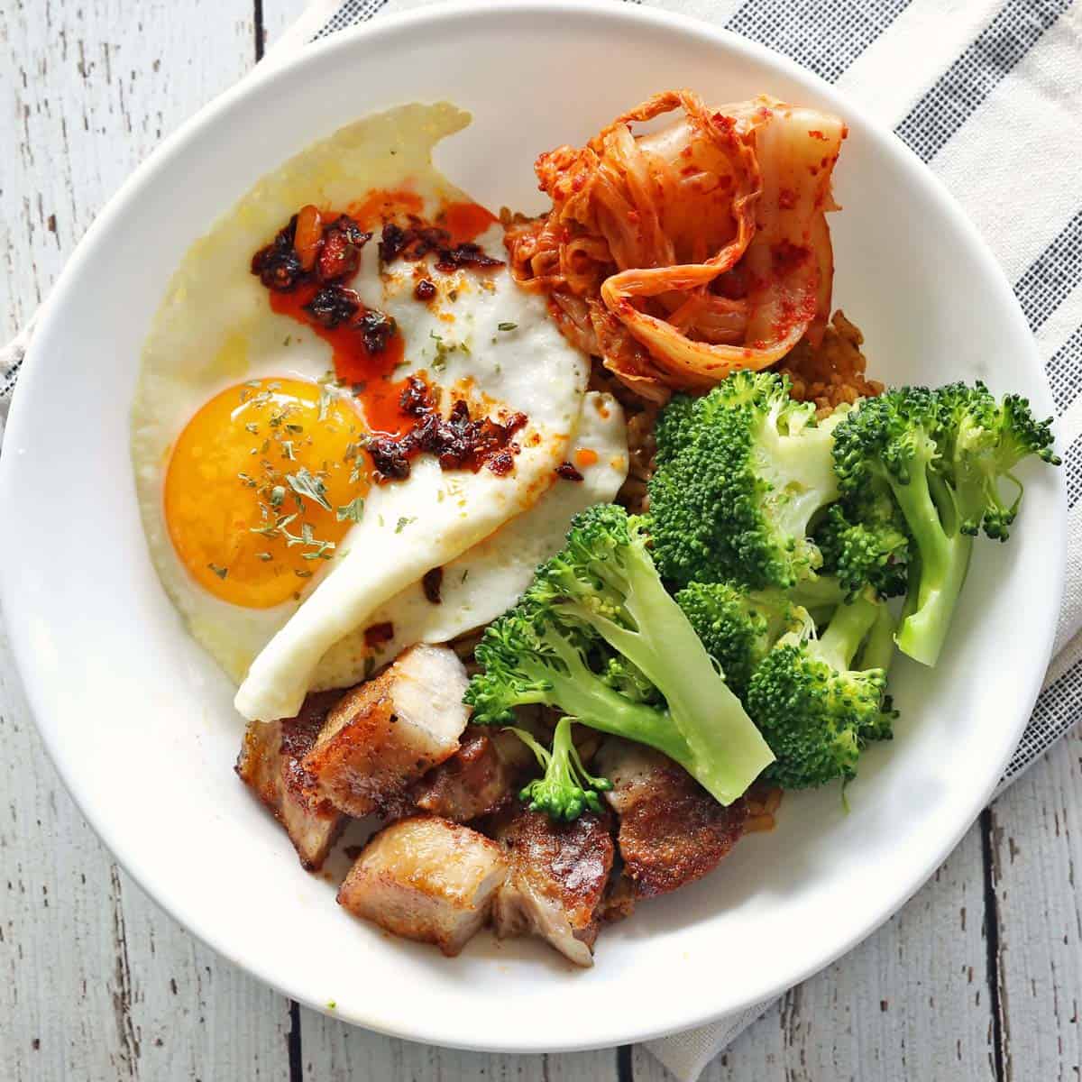 A bowl with pork belly, broccoli, a fried egg, and kimchi.