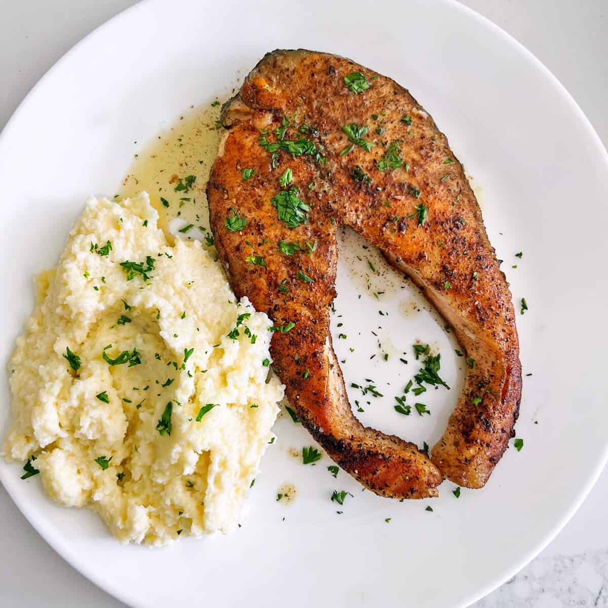 A salmon steak is served on a white plate with mashed cauliflower.