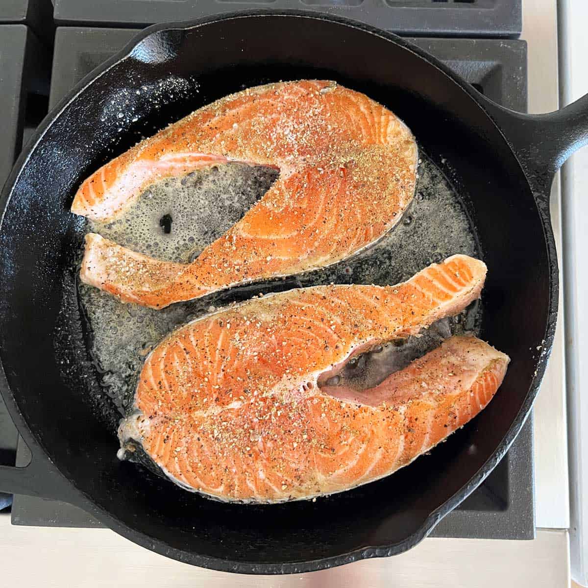 Two salmon steaks are cooking in a cast-iron skillet.
