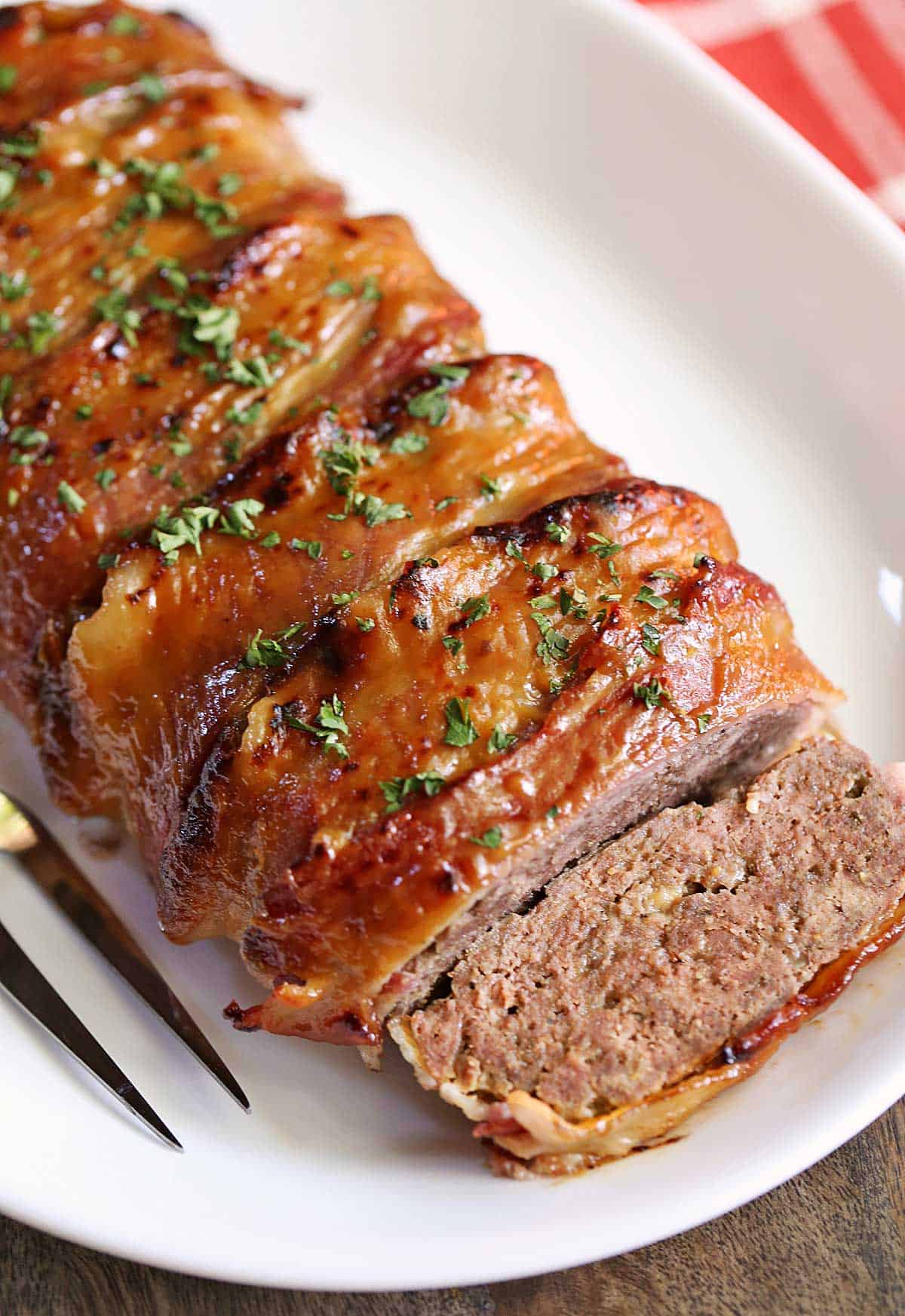 Bacon-wrapped meatloaf is served on a white platter with a serving fork.