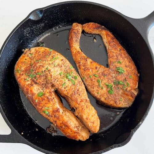 Two salmon steaks in a skillet are garnished with chopped parsley.