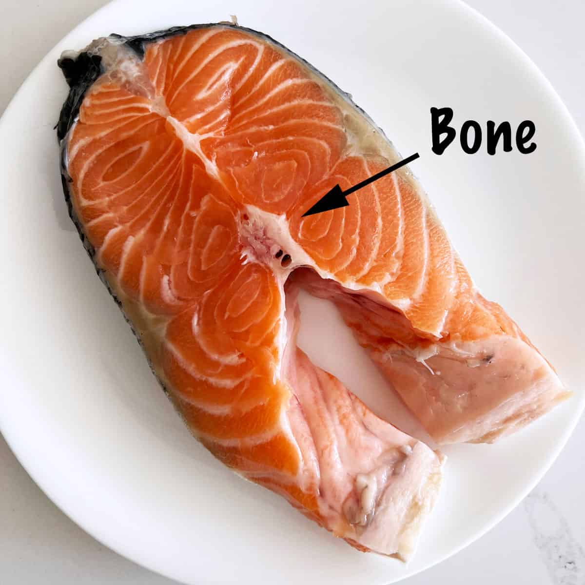 A raw salmon steak on a plate. An arrow and a text point out to where the bone is located.