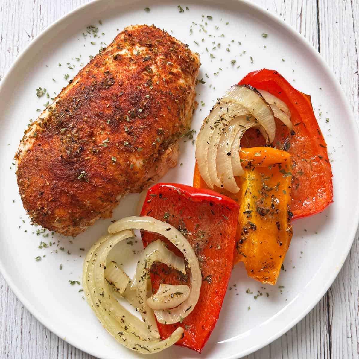 A baked chicken breast is served on a white plate with roasted peppers and onions.