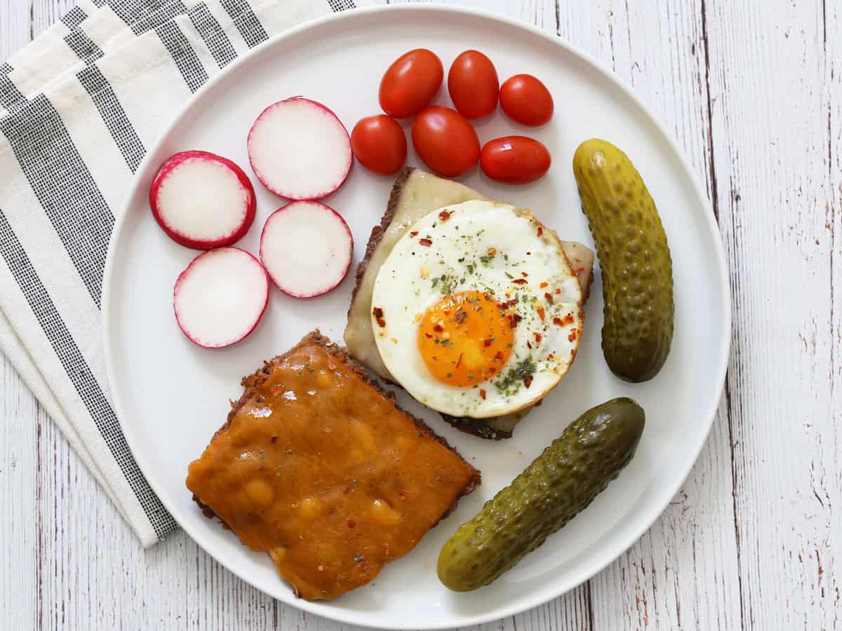 Two baked hamburgers are served with a fried egg, veggies, and pickles.