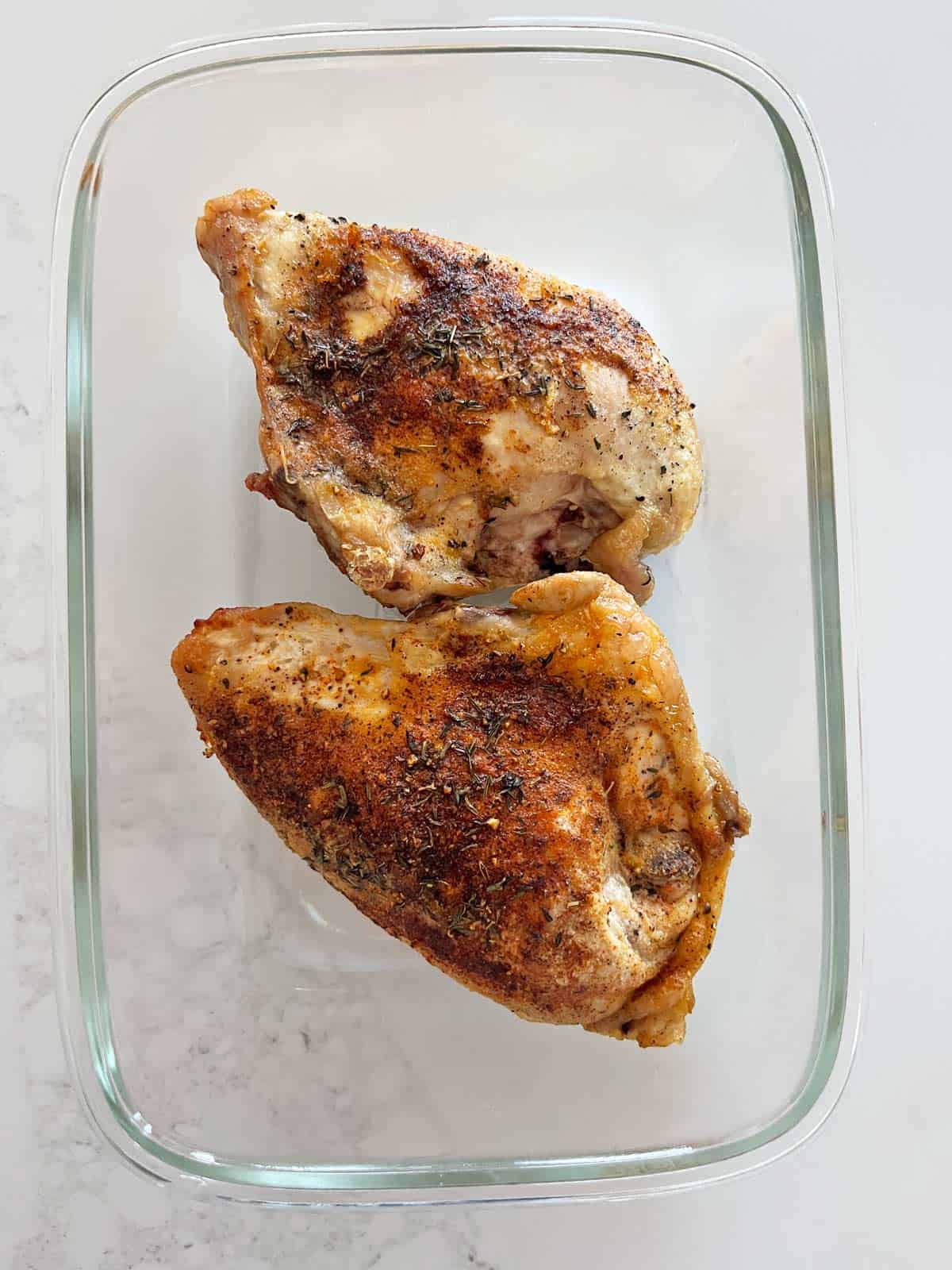 Two skin-on, bone-in chicken breasts are stored in a glass food storage containers.