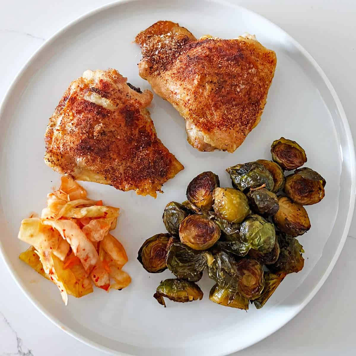 Two baked chicken thighs are served with roasted Brussels sprouts and kimchi.
