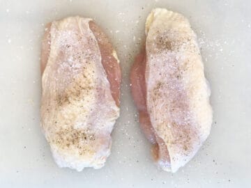 Two skin-on chicken breasts are seasoned with salt and pepper.