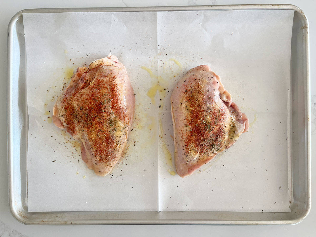 Two seasoned skin-on, bone-in chicken breasts on a rimmed baking sheet.