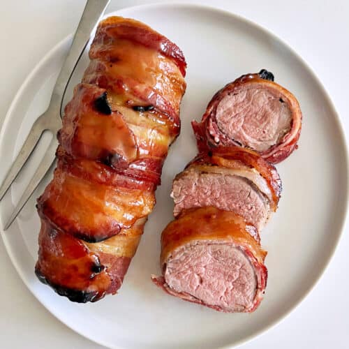 Bacon-wrapped pork tenderloin is served on a white plate with a serving fork.