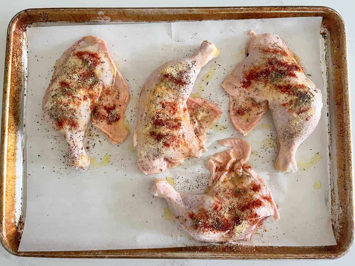 Four chicken leg quarters on a baking sheet, seasoned with salt and spices.
