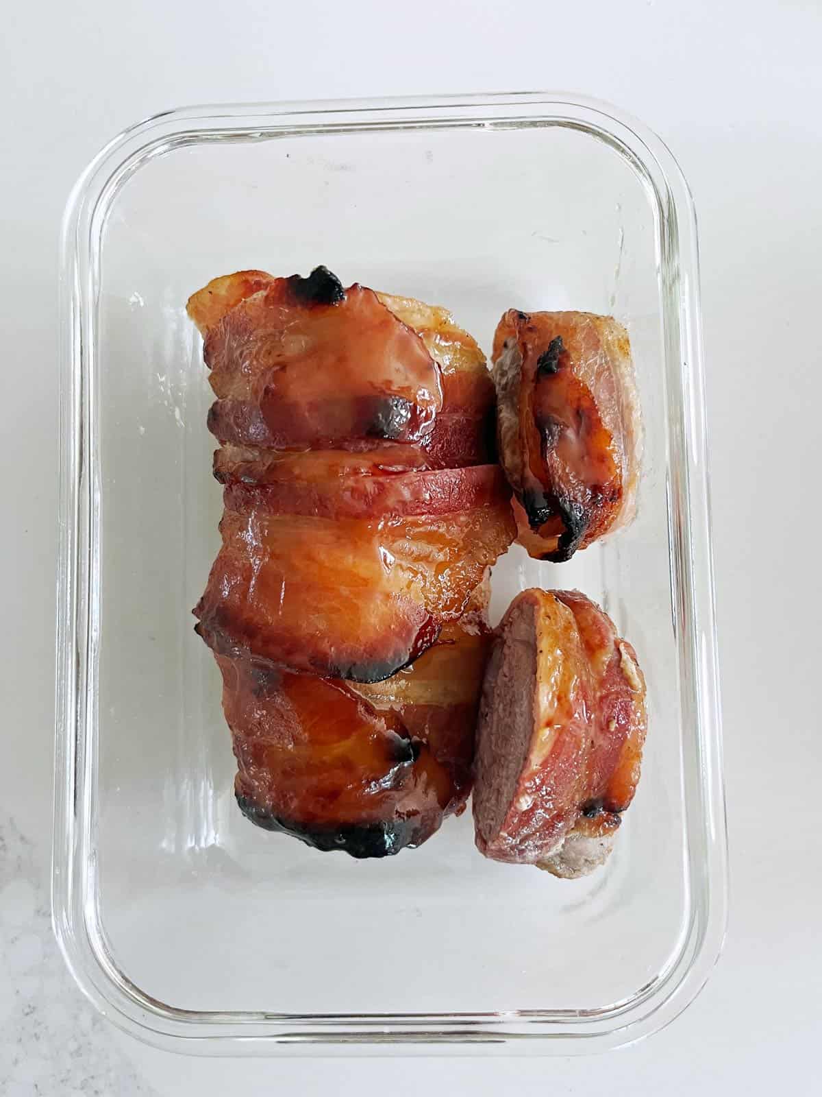 Bacon-wrapped pork tenderloin leftovers are kept in a glass food storage container.