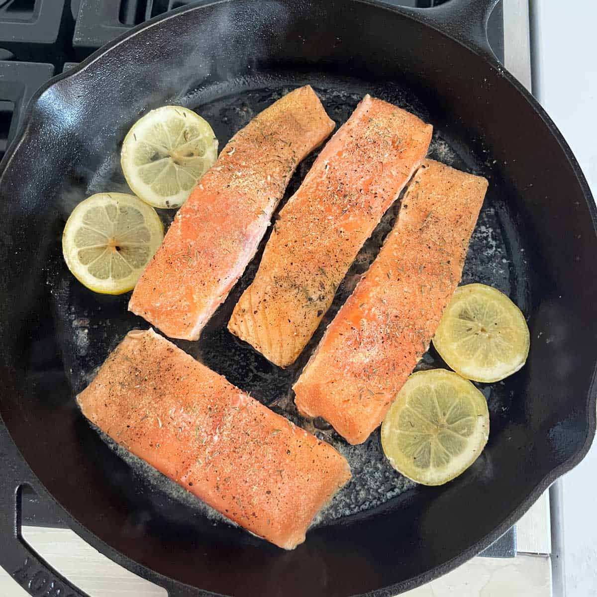 Four salmon fillets and four lemon slices are seared in a cast-iron skillet.