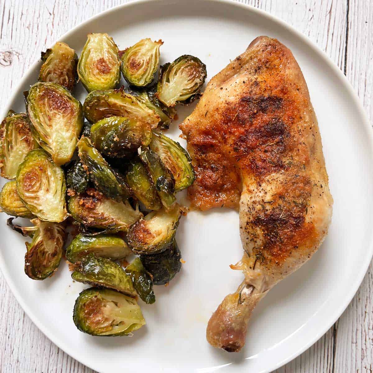 A chicken quarter is served on a white plate with roasted Brussels sprouts.